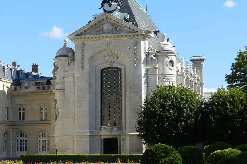 Façade claire d'une chapelle à fronton surmonté d'une statue, au fond d'un jardin à la française, sous un ciel bleu